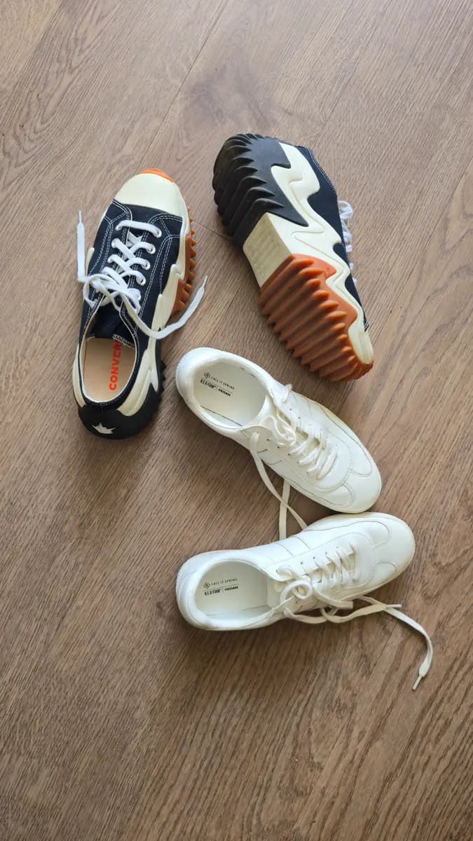 Flat-lay of sneakers on a light wood floor — a black-and-cream low-top and a chunky orange-soled trainer beside a pair of white leather sneakers