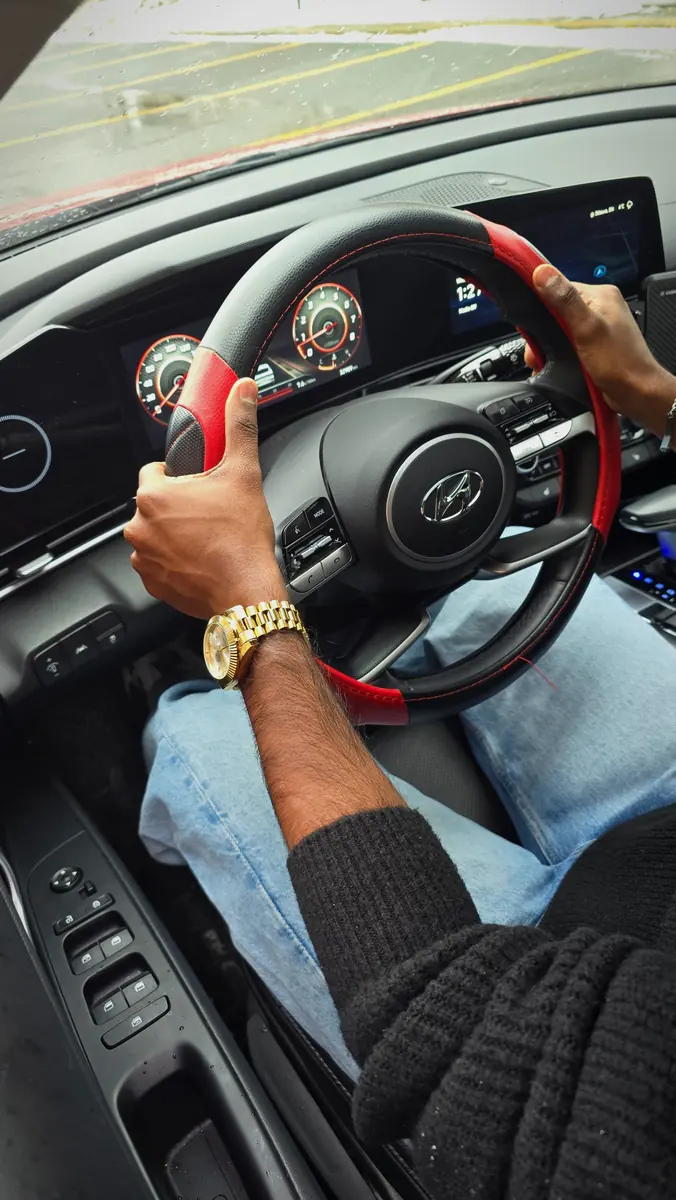 Driver's-eye view of hands on a red-and-black steering wheel, a gold watch on the wrist