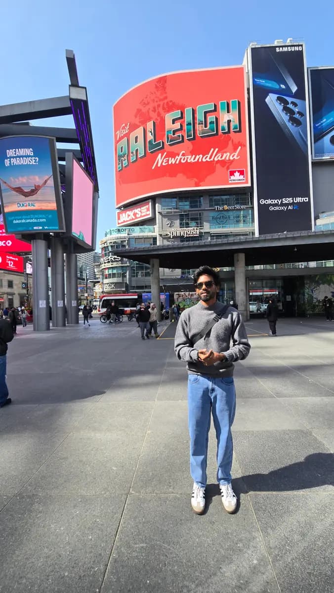 Vijay standing in Yonge-Dundas Square in Toronto, surrounded by billboards under a clear blue sky